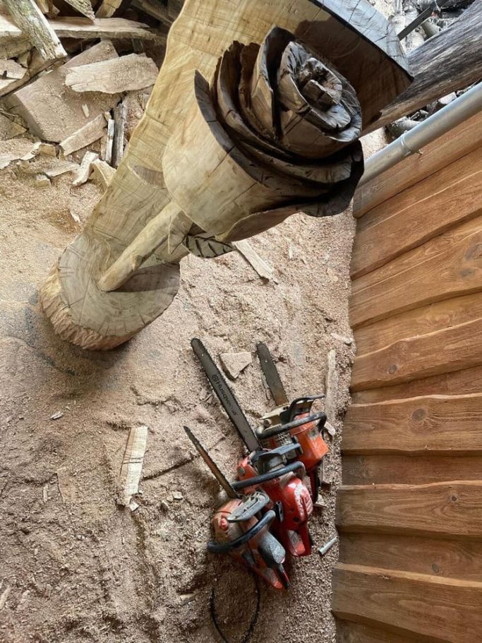 Geschnitzte Rose auf einem Holzstamm im Hintergrund liegen drei Motorsägen auf dem Boden