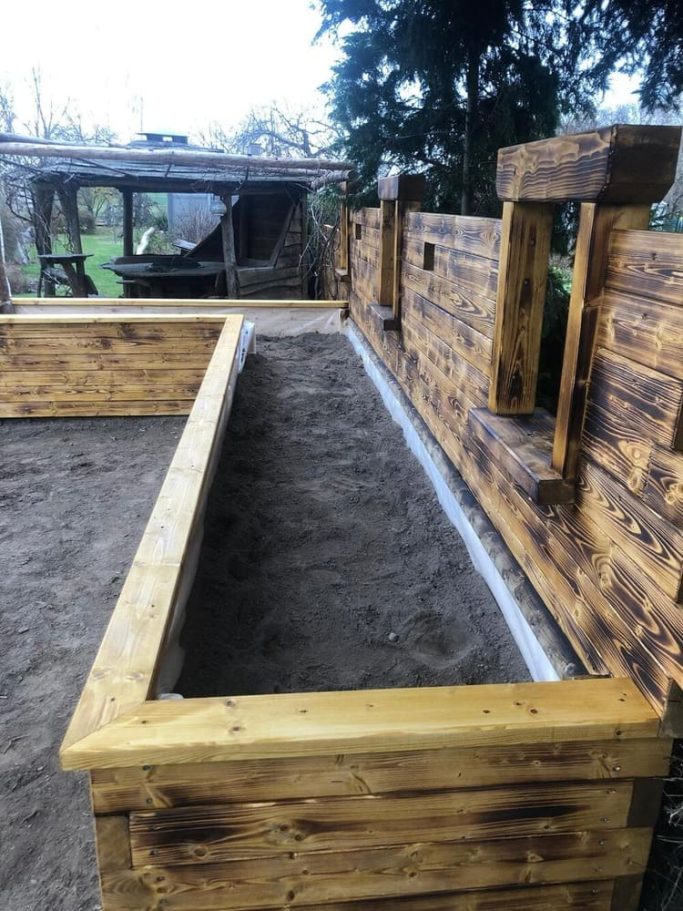 Hochbeet in einem Holzkasten mit Sand, umgeben von einer Holztrennwand und Bäumen im Hintergrund.